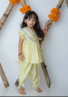 A young girl wearing a traditional dhoti set with embroidery, posing against a backdrop of bamboo and decorative flowers.