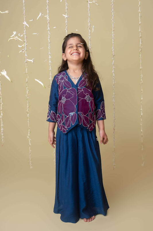 A young girl wearing a traditional midnight gold applique sharara jacket set with zari threadwork and sequin pearl accents, standing against a neutral backdrop.