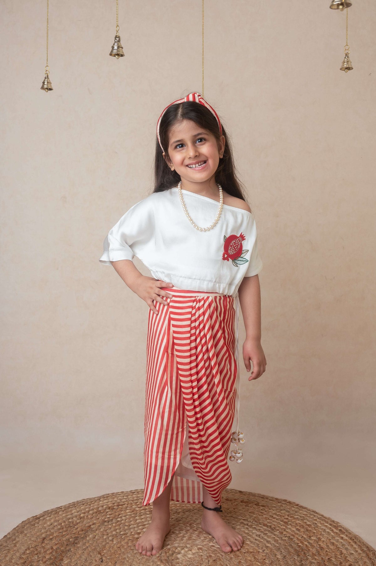 A child model wearing an off shoulder top with drawstring waist and tassels, paired with a custom printed draped skirt. The outfit is primarily white and red.