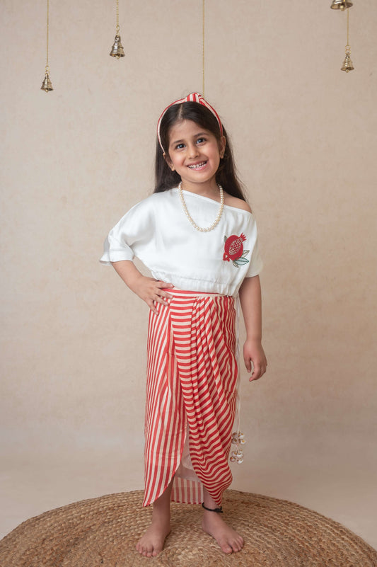A child model wearing an off shoulder top with drawstring waist and tassels, paired with a custom printed draped skirt. The outfit is primarily white and red.