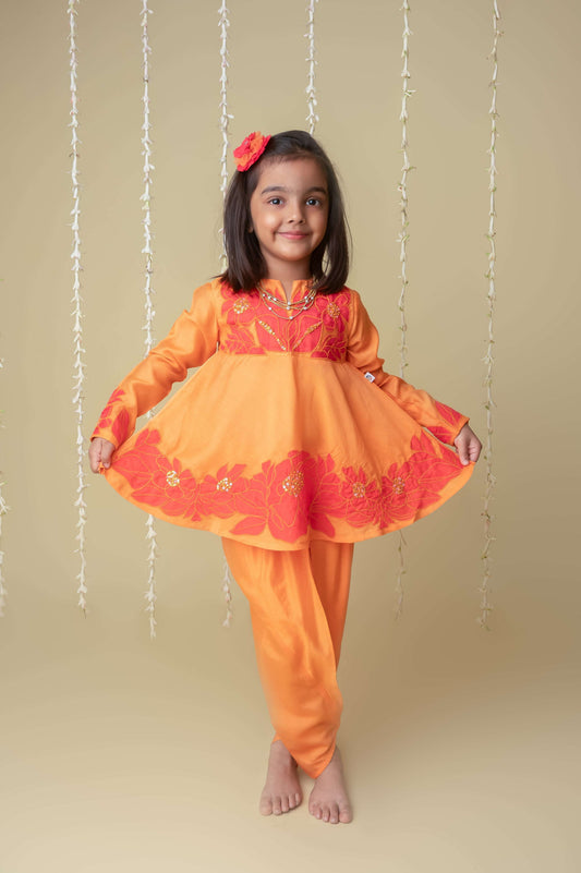 A young girl wearing a traditional Indian outfit consisting of an orange and red blouse with floral patterns and matching dhoti pants, standing against a plain background.