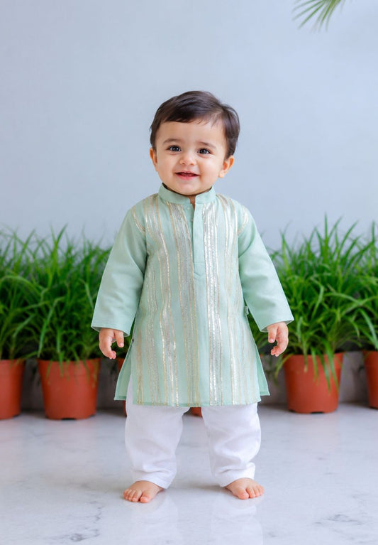 A toddler standing barefoot indoors, wearing a light mint green traditional tunic with vertical metallic gold stripes and white pants. The background features a light gray wall, potted green plants, and a white marble floor.